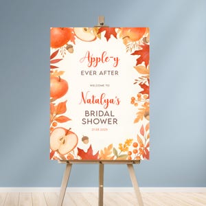 Puede incluir: Un letrero de bienvenida para una despedida de soltera, exhibido en un caballete de madera. El letrero presenta el texto "Apple-y Ever After" y "Natalya's Bridal Shower" con un tema otoñal. El diseño incluye ilustraciones de acuarela de manzanas, hojas y bellotas.