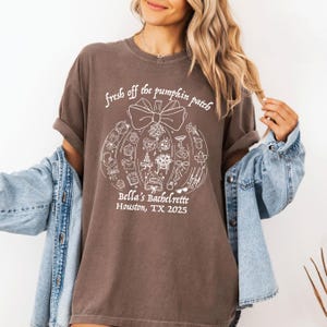 Może przedstawiać: Brązowa koszulka z napisem "fresh off the pumpkin patch" i grafiką dyni. Grafika zawiera różne ikony oraz napis "Bella's Bachelorette Houston, TX 2025". Koszulka ma podwinięte rękawy.