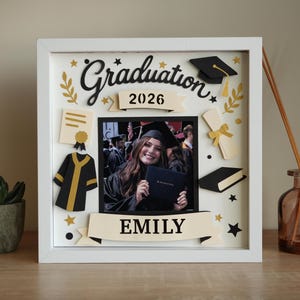 May include: A white shadow box frame containing a central photograph of a graduate holding a diploma. The frame features layered paper cutouts including a graduation cap, a gown, a diploma, and the text Graduation, 2026, and EMILY.