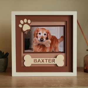 May include: A square white picture frame featuring a layered brown mat with a cutout paw print in the top left corner and a bone-shaped cutout at the bottom containing the name BAXTER. The center displays a photo of a golden dog holding a toy.