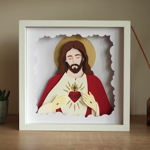 May include: A layered paper art shadow box featuring a depiction of the Sacred Heart of Jesus. The figure has brown hair, a beard, and a gold halo, wearing a red robe over a white garment with a central heart emblem featuring a cross and thorns.