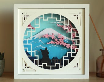 Puede incluir: Una obra de arte en papel 3D enmarcada en blanco que representa el Monte Fuji con una silueta de gato negro. La escena incluye flores de cerezo rosas y un cielo azul, enmarcado por un borde blanco decorativo.