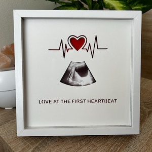 May include: White framed print featuring a red heart and heartbeat line design, an ultrasound image, and the text "LOVE AT THE FIRST HEARTBEAT". The frame is square and rests on a wooden surface.