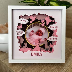 Op de afbeelding: Een wit omkaderd papieren kunstwerk met een pasgeboren baby in roze kleding. Het kunstwerk bevat de naam van de baby, de geboortetijd, de datum, het gewicht (3,5 kg) en de lengte (51 cm). Roze wolken, vlinders en sterren omringen de baby.