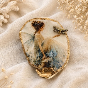 May include: An irregular, shell-shaped decorative dish with a gold-painted edge. The center features a printed illustration of a seated figure with wings, wearing a flowing blue and white gown. A small, metallic bronze-colored dragonfly is attached.