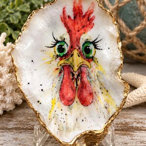 May include: A decorative oyster shell featuring a hand-painted illustration of a chicken face with bright green eyes, a red comb, and red wattles. The shell has a natural white interior and a textured, metallic gold-painted edge.