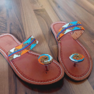 Könnte beinhalten: Ein Paar hellbraune Ledersandalen mit Zehensteg und einem breiten Riemen über dem Fuß. Der Riemen und der runde Zehensteg sind mit aufwendigen Perlenstickereien in leuchtenden Mustern aus orangefarbenen, türkisfarbenen, weißen und goldfarbenen Perlen verziert.