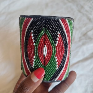 Könnte beinhalten: Ein Armband mit Perlenbesatz mit einem Muster aus roten, schwarzen, grünen und weißen Perlen. Das Armband hat eine schwarze Lederunterseite und eine zylindrische Form. Das Design ist mit einem ovalen Muster zentriert.
