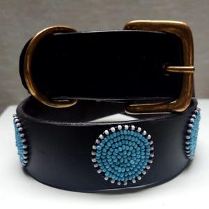 Könnte beinhalten: Schwarzer Ledergürtel mit einer Messingschnalle. Der Gürtel ist mit türkisfarbenen und weißen Perlenmustern verziert. Die Schnalle hat eine rechteckige Form mit abgerundeten Kanten.