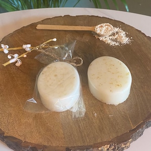 May include: Two round white soap bars with gold flecks on a wooden surface. The soap bars are wrapped in clear plastic and tied with twine.