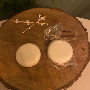 May include: Two circular, white soaps, one in clear cellophane tied with twine, are displayed on a rustic wooden slab. A small decorative twig with white details is also present. The soaps have a smooth, clean appearance.