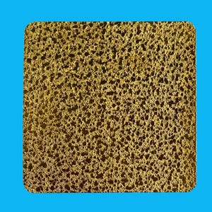 May include: A square, gold-coloured textured surface with rounded corners, set against a bright blue background. The surface has a porous, sponge-like appearance, with numerous small holes and a metallic sheen. The overall effect is of a decorative or artistic element.