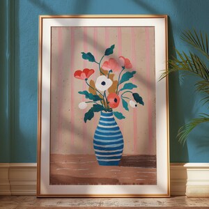 May include: A framed art print featuring a still life of flowers in a blue and white striped vase. The flowers are red, pink, and white with green leaves. The background has pink vertical stripes. The frame is gold.