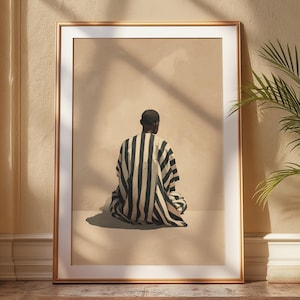 May include: A framed art print showing a person in a black and white striped garment, seated with their back to the viewer. The artwork is set against a neutral background, framed in gold, and displayed in a room with natural light.