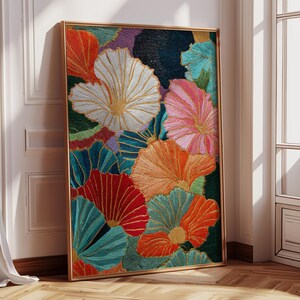 May include: A framed floral artwork featuring embroidered flowers in shades of red, orange, teal, pink, and white. The artwork has a gold frame and is leaning against a white wall near a window.
