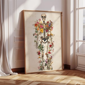 May include: A framed print featuring a human skeleton adorned with colorful flowers. The artwork has a vintage aesthetic, with the skeleton's bones and the flowers' vibrant hues contrasting against a cream-colored background. The frame is a warm gold color.