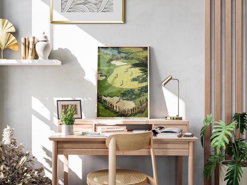 K&ouml;nnte beinhalten: Ein gerahmter Golfplatzdruck in Gr&uuml;n- und Gelbt&ouml;nen, der auf einem Holzschreibtisch ausgestellt ist. Der Druck zeigt Golfer auf einem Platz mit Zuschauern. Ein kleines gerahmtes Bild und eine Topfpflanze befinden sich ebenfalls auf dem Schreibtisch.