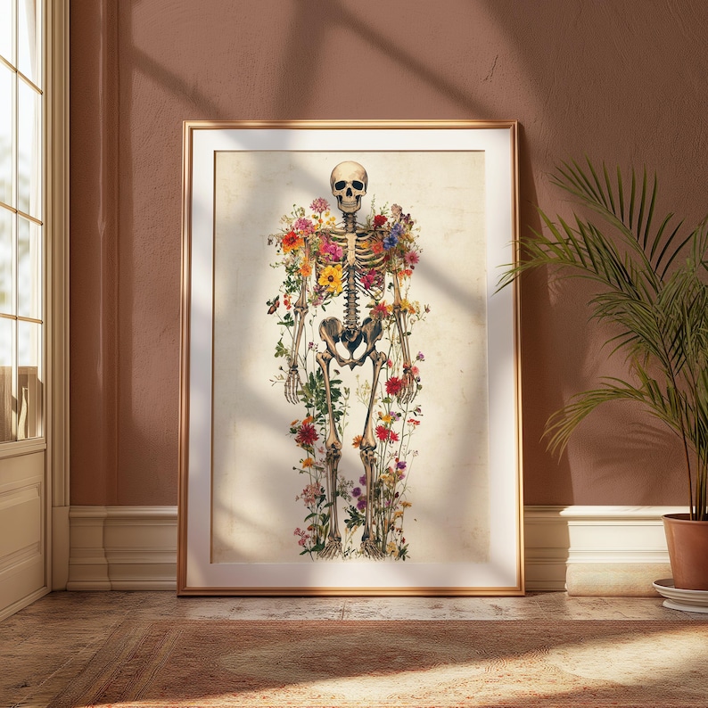 May include: A framed art print featuring a human skeleton adorned with colorful flowers. The artwork is set against a neutral background and framed in gold, creating a striking contrast. The print is displayed on a wooden floor.