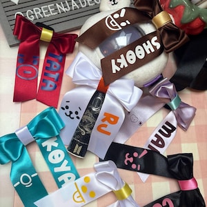 Può includere: Una collezione di nastri di raso colorati con fiocchi, ognuno con un nome o un disegno di personaggio diverso. I nastri sono in vari colori, tra cui rosso, verde acqua, bianco e nero, con accenti dorati e rosa. Alcuni nastri hanno del testo.