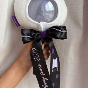 Può includere: Un bastoncino luminoso in peluche bianco con un globo trasparente, nastro nero e dettagli viola. Il nastro riporta la scritta "Army since 2019" e il logo BTS. Il bastoncino luminoso è a forma di orso.