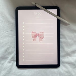 May include: A black tablet displaying a pink to-do list with a bow illustration and the words "TO DO list". A white stylus rests on the tablet. The tablet is on a white surface.