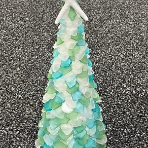 Puede incluir: Un árbol de Navidad decorativo hecho de vidrio de mar en tonos blanco, verde y turquesa. El árbol está rematado con una estrella de mar blanca. Las piezas de vidrio de mar están dispuestas para parecerse a escamas, creando un adorno único con temática costera.