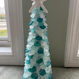 Puede incluir: Escultura decorativa en forma de árbol hecha de vidrio marino turquesa y blanco, coronada por una estrella de mar blanca. El árbol mide unos 30 cm de altura, ideal para decoración costera.