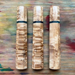 May include: Three wooden knife handles with a light brown, patterned finish. Each handle has a black band near the top. The handles are arranged vertically on a multicolored wooden surface.