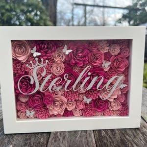 May include: A white frame with a pink and white paper flower arrangement inside. The name "Sterling" is written in silver glitter on the flowers.