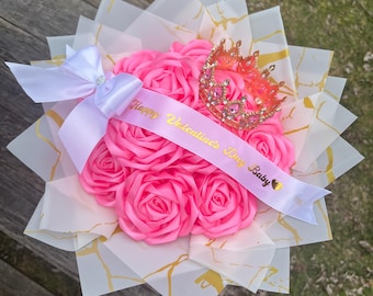 May include: A bouquet of pink roses wrapped in white and gold paper, adorned with a pink ribbon, a gold crown, and a sash that reads "Happy Valentine's Day Baby."