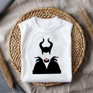 Puede incluir: Camiseta blanca con una silueta negra de Mal&eacute;fica de la pel&iacute;cula de Disney La Bella Durmiente. La silueta incluye sus cuernos, cabello y vestido.