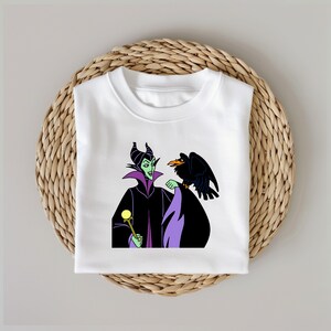 Puede incluir: Camiseta blanca con un estampado gr&aacute;fico de Mal&eacute;fica de La Bella Durmiente de Disney. Mal&eacute;fica lleva un vestido morado y negro y sostiene un bast&oacute;n dorado. Un cuervo negro est&aacute; posado en su hombro.