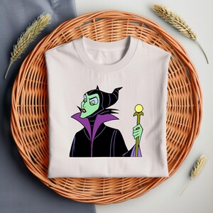 Puede incluir: Camiseta blanca con una imagen de dibujos animados de Mal&eacute;fica de La Bella Durmiente de Disney. Lleva un vestido negro y morado y sostiene un bast&oacute;n con un orbe amarillo en la parte superior.