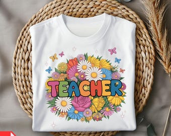 Könnte beinhalten: Weißes T-Shirt mit einem farbenfrohen Blumendesign und dem Wort "TEACHER" in Regenbogenbuchstaben. Das Design beinhaltet Gänseblümchen, Rosen und Schmetterlinge. Das T-Shirt liegt auf einer gewebten Oberfläche.