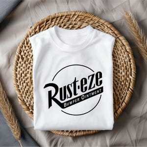 Puede incluir: Camiseta blanca con un gr&aacute;fico circular negro que dice "Rust-eze Bumper Ointment" en una fuente de estilo vintage.