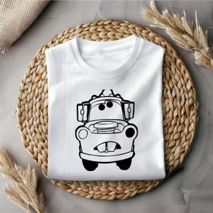Puede incluir: Una camiseta blanca con una ilustraci&oacute;n en blanco y negro de Mater de la pel&iacute;cula Cars de Disney Pixar. Mater es una gr&uacute;a con una gran sonrisa y ojos grandes.