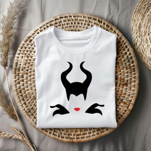 Puede incluir: Camiseta blanca con una silueta negra de la cara y los cuernos de Mal&eacute;fica. La silueta tiene labios rojos.