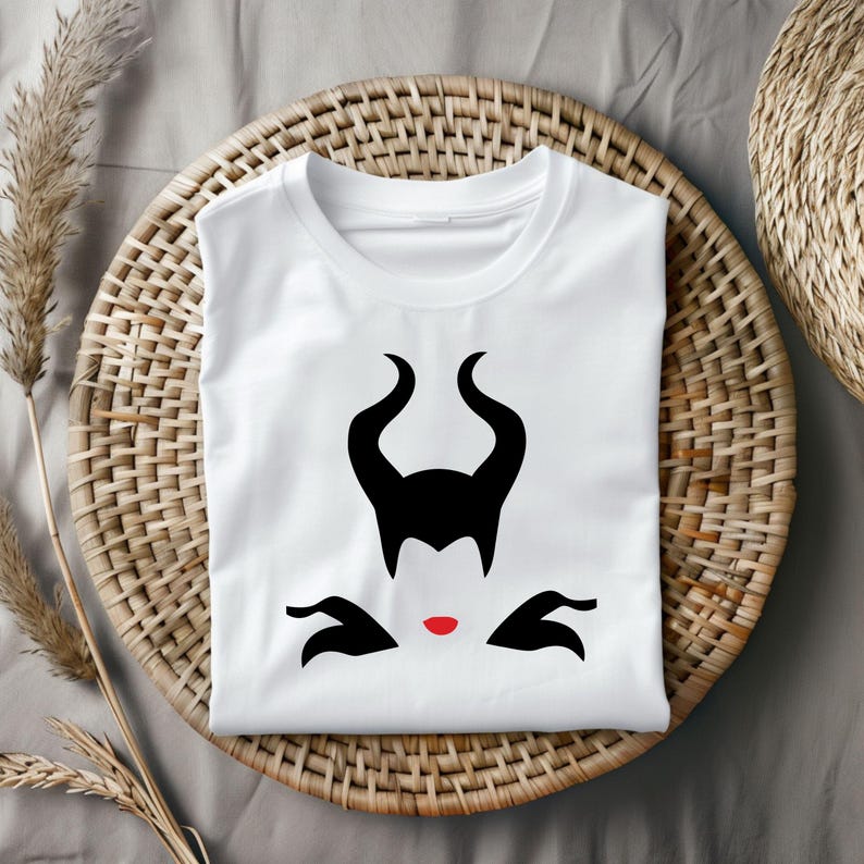 Puede incluir: Camiseta blanca con una silueta negra de la cara y los cuernos de Mal&eacute;fica. La silueta tiene labios rojos.