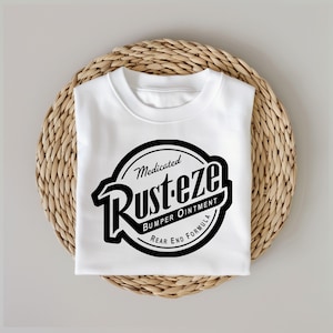 Puede incluir: Camiseta blanca con un gr&aacute;fico en blanco y negro de un c&iacute;rculo con el texto "Medicated Rust-eze Bumper Ointment Rear End Formula" en su interior.