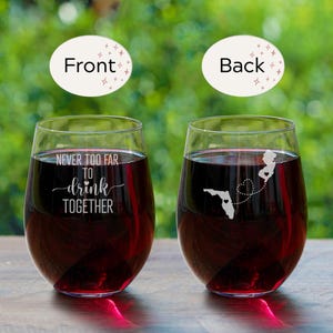 Puede incluir: Dos copas de vino transparentes con vino tinto. Una copa tiene el texto "Never too far to drink together" grabado en la parte delantera. La otra copa tiene un contorno de Florida y Nueva Jersey con un coraz&oacute;n que los conecta, grabado en la parte posterior.