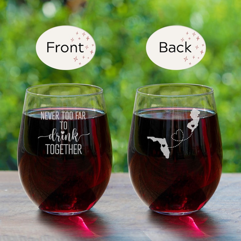 Puede incluir: Dos copas de vino transparentes con vino tinto. Una copa tiene el texto "Never too far to drink together" grabado en la parte delantera. La otra copa tiene un contorno de Florida y Nueva Jersey con un coraz&oacute;n que los conecta, grabado en la parte posterior.