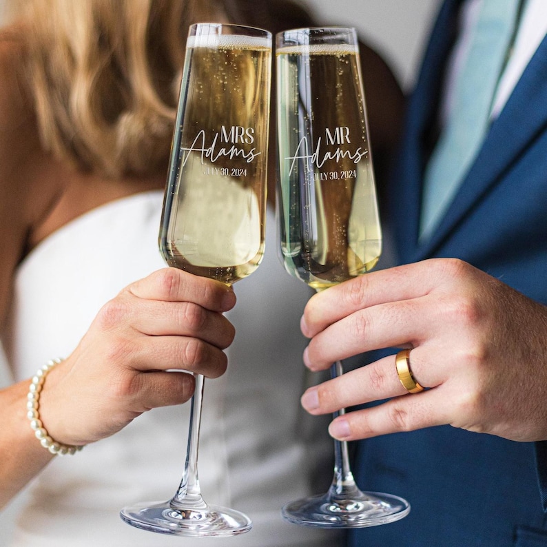 May include: Two champagne flutes filled with a bubbly beverage. Each glass is etched with the names "Mrs. Adams" and "Mr. Adams" along with the date "July 30, 2024". A gold wedding band is visible on a finger.