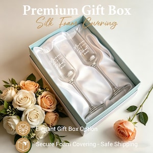May include: A gift box with two champagne flutes, personalized with names and a date. The flutes rest on a white satin lining within a light blue box. The box is surrounded by a bouquet of roses and a single rose.