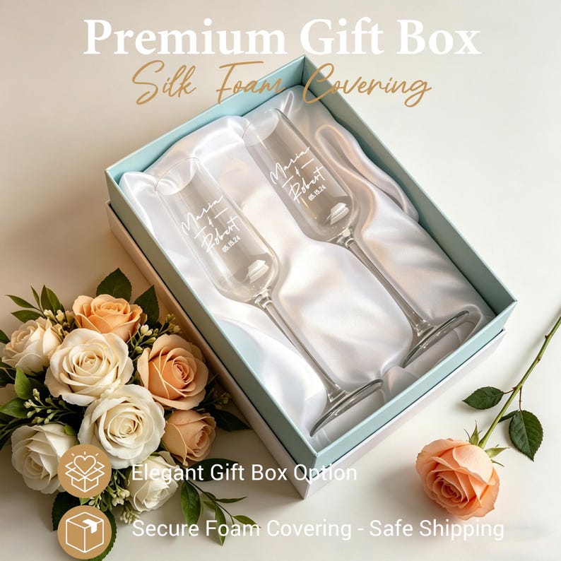 May include: A gift box with two champagne flutes, personalized with names and a date. The flutes rest on a white satin lining within a light blue box. The box is surrounded by a bouquet of roses and a single rose.