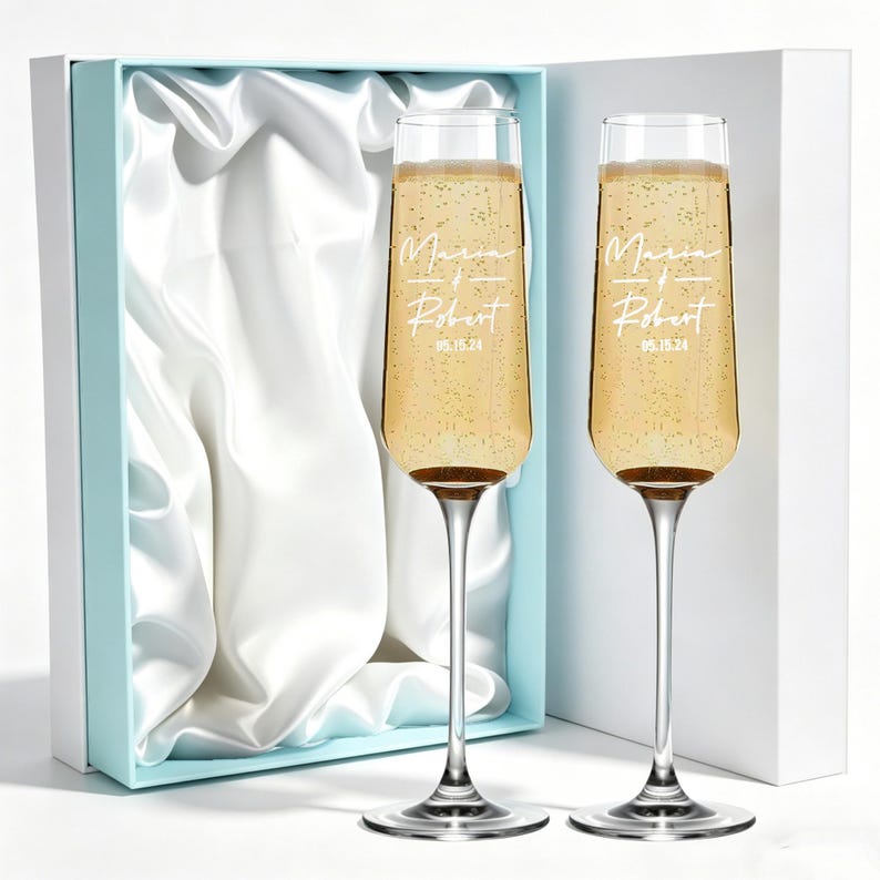 May include: Two champagne flutes filled with a bubbly beverage, each engraved with the names "Maria" and "Robert" and the date "05.15.24". The flutes sit in front of a light blue box with a white satin lining.