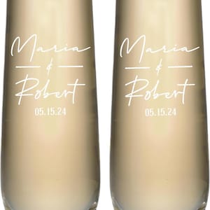 May include: Two clear champagne flutes filled with a golden liquid. Each glass is inscribed with the names "Maria & Robert" and the date "05.15.24" in white script. The flutes have a slender, elegant design, perfect for a wedding or celebration.