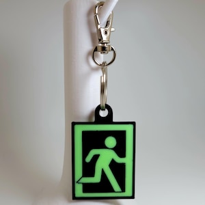 May include: A black and green glow-in-the-dark keychain with a running person icon inside a doorway. The keychain has a silver metal ring and a silver metal clip.
