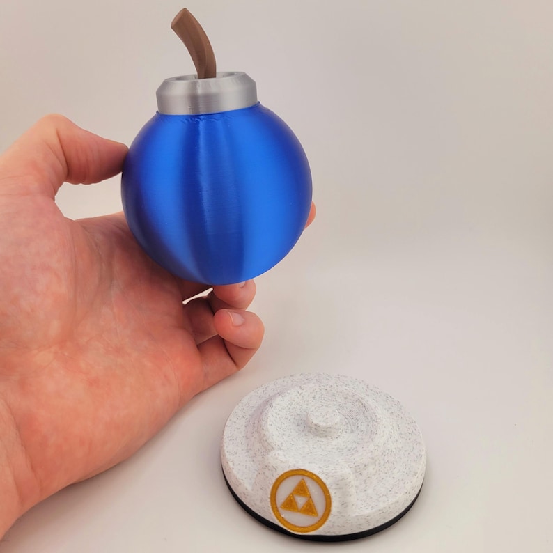 Op de afbeelding: Een blauw, bolvormig rekwisiet met een bruine steel en zilveren accenten, boven een getextureerde witte basis met een gouden embleem. De prop lijkt op een bom uit een videogame.
