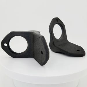 May include: Two black plastic wall mount brackets with a circular hole in the center. The brackets are designed to hold a round object, such as a pipe or a speaker.