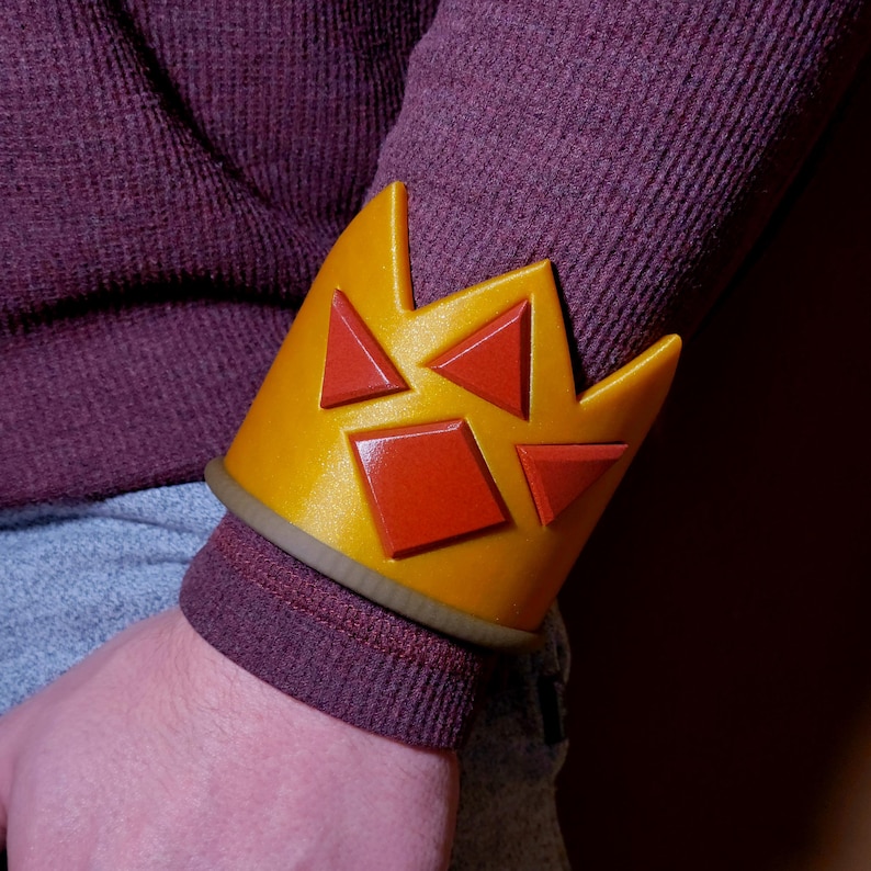 Puede incluir: Un brazalete de color amarillo y rojo con forma de corona y un dise&ntilde;o geom&eacute;trico. El brazalete se lleva en la mu&ntilde;eca.